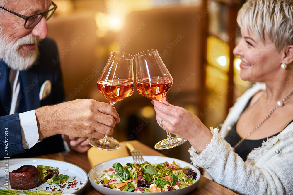 Beautiful elderly couple have a meal in personable restaurant, in love. Grey-haired male and female drink and clink glasses with champagne. Beautiful elderly couple have a meal in personable restaurant, in love. Grey-haired male and female drink and clink glasses with champagne.