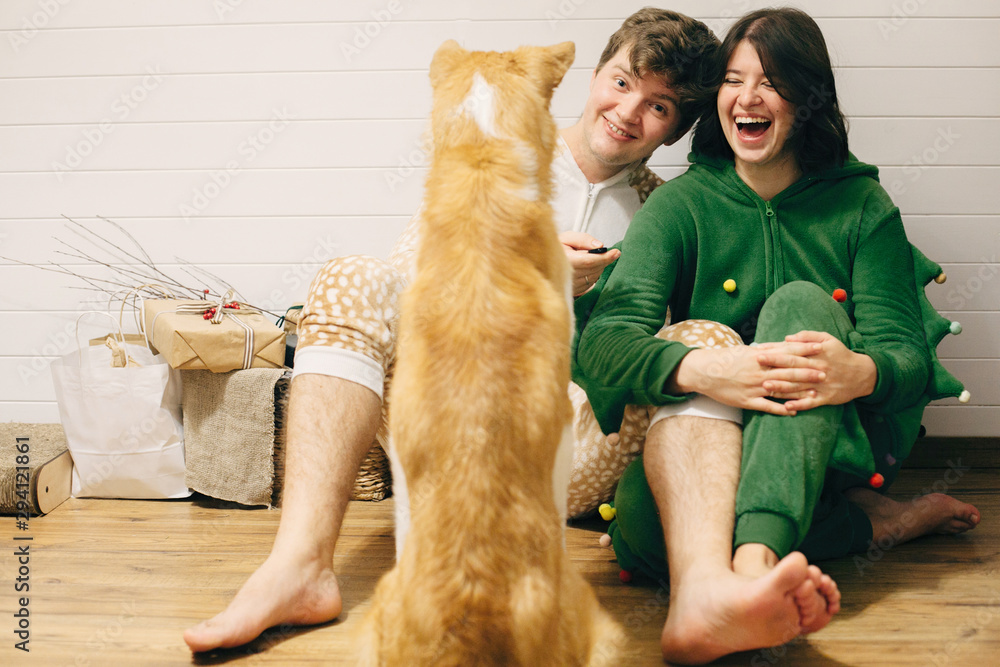 Happy couple in festive pajamas having fun and feeding their dog with canned food in room. Celebrating Christmas or New Year eve together with pets. Happy couple in festive pajamas having fun and feeding their dog with canned food in room. Celebrating Christmas or New Year eve together with pets.
