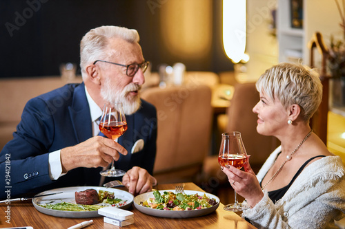 Stylish, elegant senior couple look at each other and chatting, close relationship. Grey-haired man in tuxedo raise a glass. Use e-cigarette, vape. Modern technologies