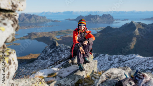young woman hiker sitting on top of the Rundfjelle mountain with views of the surrounding snowy peaks and the sea in Lofoten, Norway