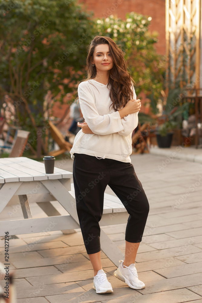 Elegant stylish girl in in white hoodie and black trousers sitting and relaxing in summer cafe at sunset. Woman enjoying cup of coffee Elegant stylish girl in in white hoodie and black trousers sitting and relaxing in summer cafe at sunset. Woman enjoying cup of coffee