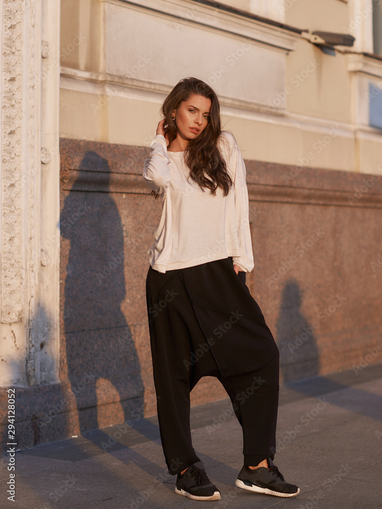 Outdoor summer portrait. Young stylish woman in white hoodie and black trousers standing and posing at city street at sunset Outdoor summer portrait. Young stylish woman in white hoodie and black trousers standing and posing at city street at sunset