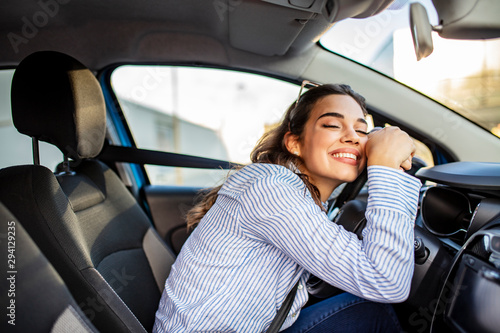 Young Woman Embracing Her New Car. Excited young woman and her new car indoors. Young and cheerful woman enjoying new car hugging steering wheel sitting inside