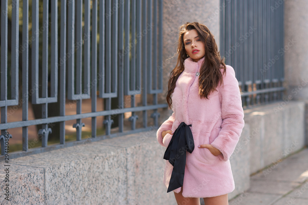 Young elegant woman in pink short vegan faux fur coat posing at city street. Fashion style outdoor portrait