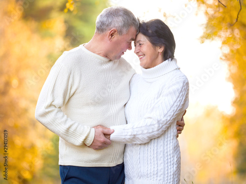Beautiful elderly couple in white knitted sweaters stand hugging in the autumn forest