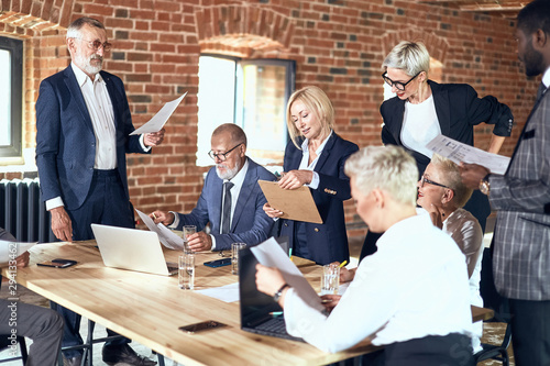 Group of businessmen in stylish suits at table in office made brick walls discuss project looking at papier. White and black laptops on table and glasses of water