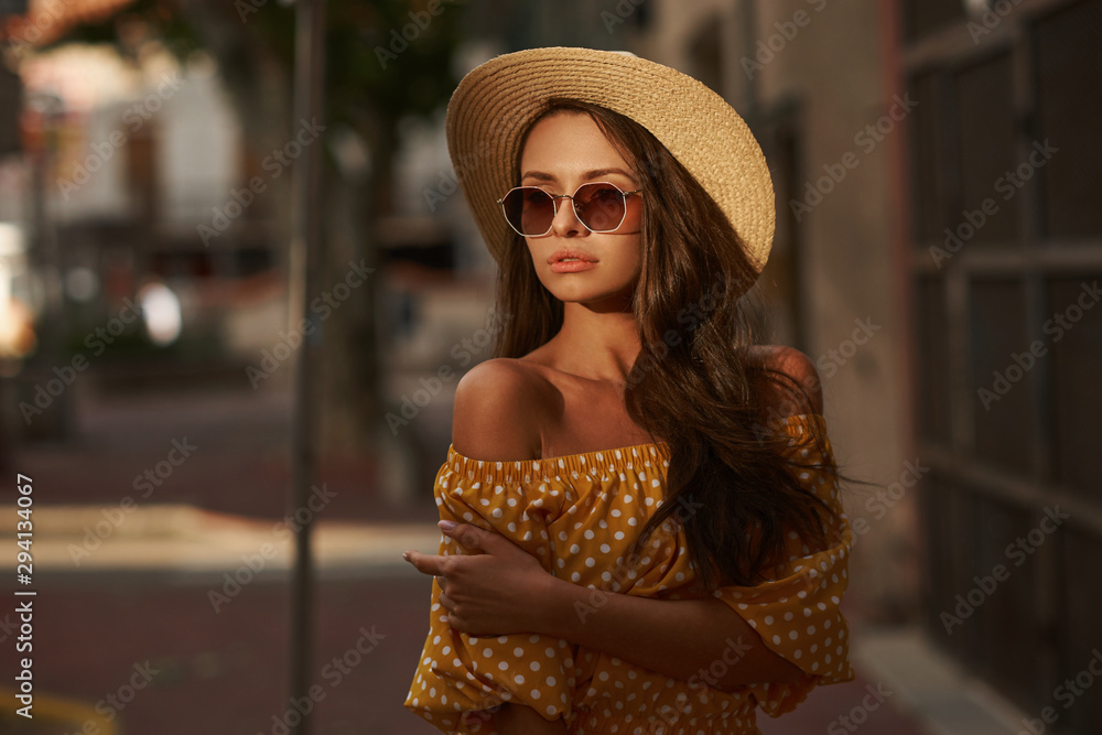Closeup outdoor portrait of young beautiful caucasian young woman with long brunette hair wearing yellow polka dot dress, sunglasses and thatch hat. Summer sunny evening street