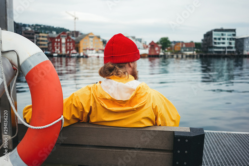 Obraz na plátně Back view of male tourist wearing yellow jacket sitting on bench in front of city pier