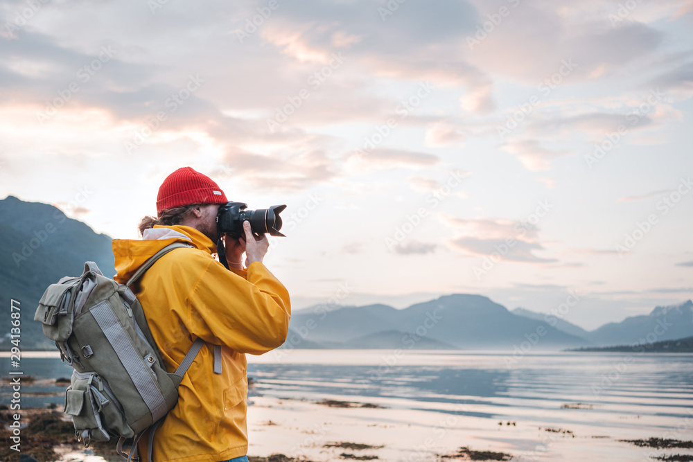 Obraz Hipster traveler photographer takes a photo beautiful nordic landscape on professional camera. Man tourist with backpack shoots a photo scandinavia nature fototapeta, plakat