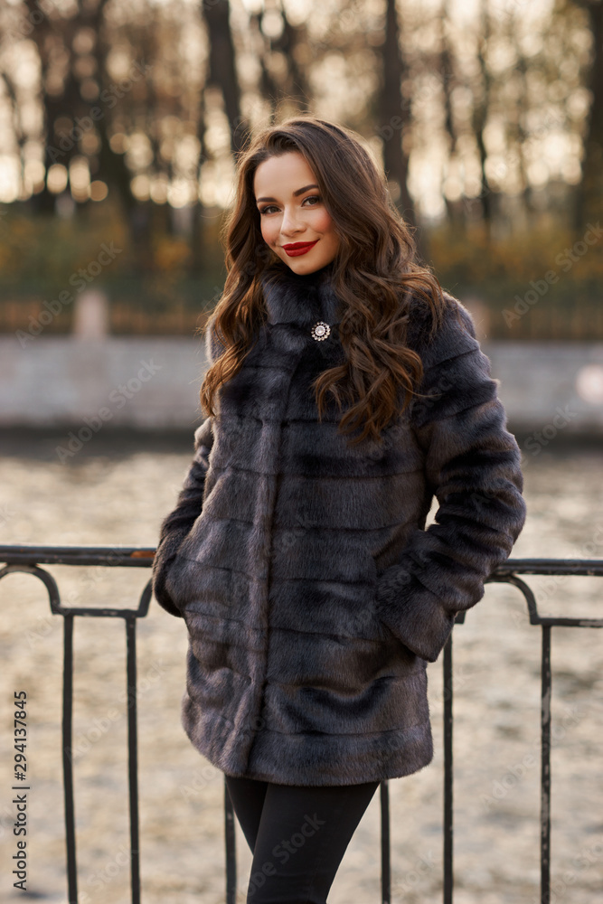 Pretty girl in short fur coat standing at river embankment and posing. Beautiful woman with long brunette wavy hair, makeup and red lips Pretty girl in short fur coat standing at river embankment and posing. Beautiful woman with long brunette wavy hair, makeup and red lips