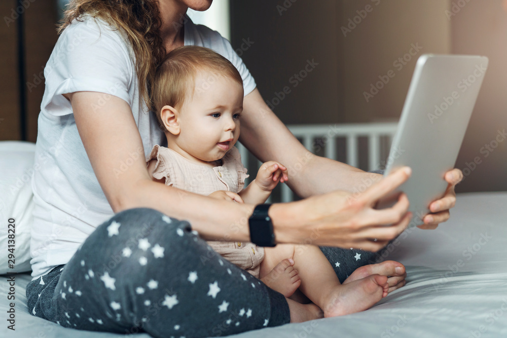 Nine-month-old baby girl with mom sitting on bed, looking at screen of digital tablet. Child development using modern technology. Nine-month-old baby girl with mom sitting on bed, looking at screen of digital tablet. Child development using modern technology.