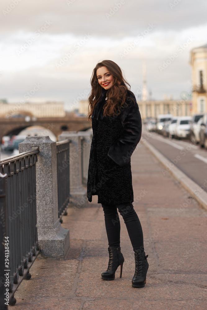 Pretty smiling woman in short black fur coat standing at river embankment in city