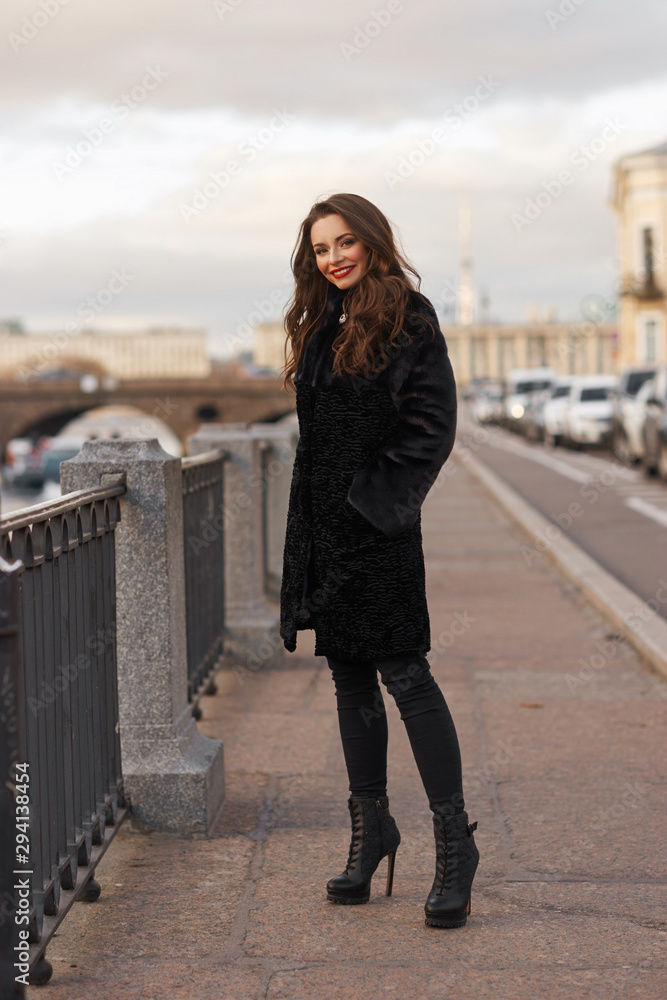 Pretty smiling woman in short black fur coat standing at river embankment in city Pretty smiling woman in short black fur coat standing at river embankment in city