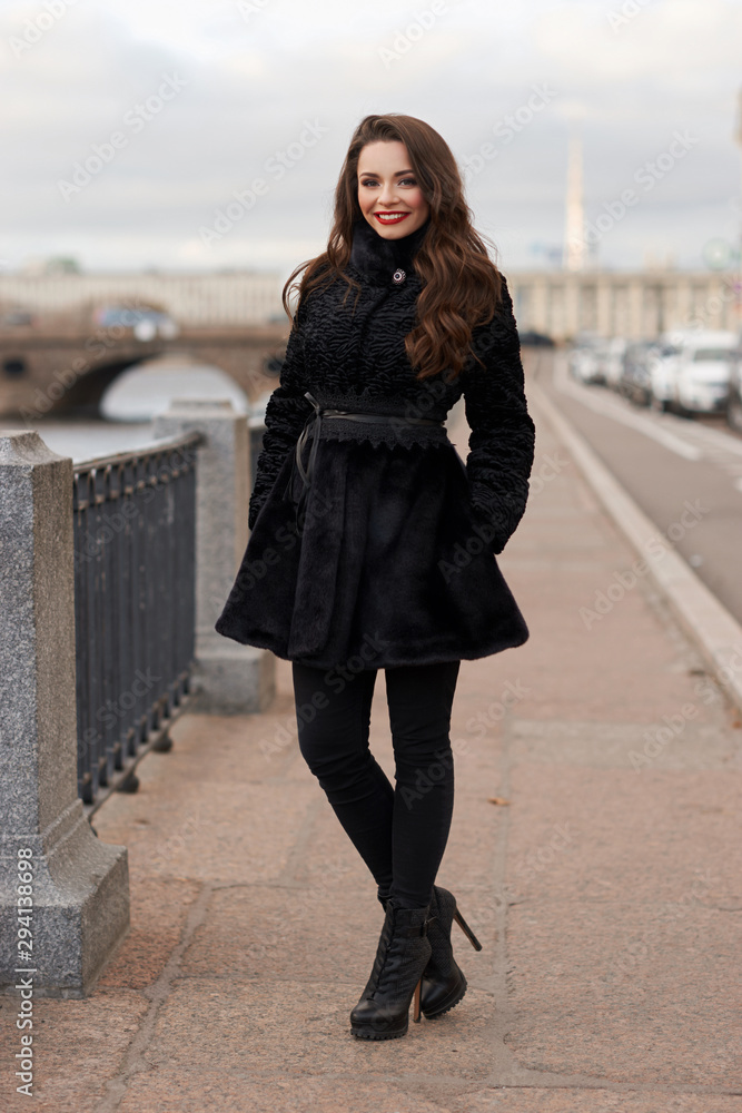 Portrait of young beautiful lady walking at street of city. Model wearing stylish black fur coat. Girl looking in camera. Female fashion concept.