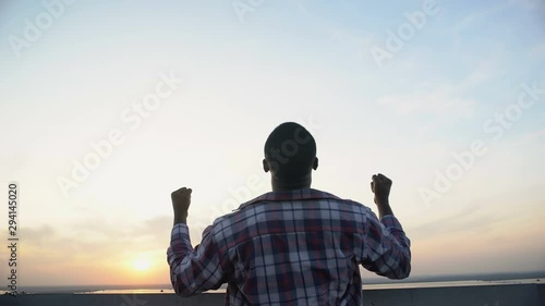 Back view of black man raising hands to sky against sunset, achievement, winner