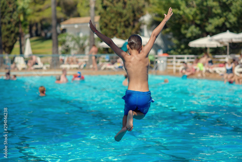 Cute active European boy in blue swimming shorts having fun in hotel   s swimming pool  he is making fantastic jump  his hands open wide.