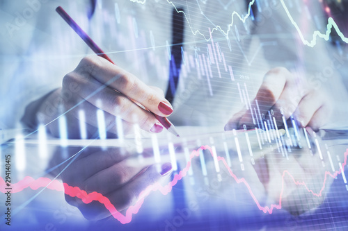 A woman hands writing information about stock market in notepad. Forex chart holograms in front. Concept of research.