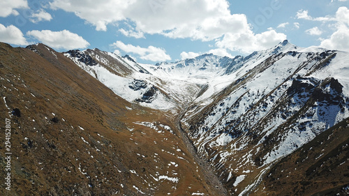 Autumn in the mountains. Yellow grass and snow-capped mountains. The gorge with the river. Blue sky and big white clouds. The stones lie on the slopes, in some places the snow. Almaty, Kazakhstan. © SergeyPanikhin