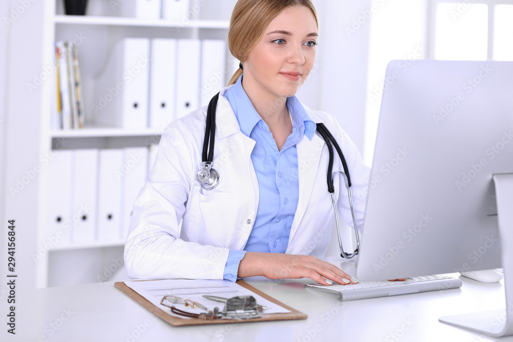 Young woman doctor at work in hospital looking at desktop pc monitor. Physician controls medication history records and exam results. Medicine and healthcare concept Young woman doctor at work in hospital looking at desktop pc monitor. Physician controls medication history records and exam results. Medicine and healthcare concept