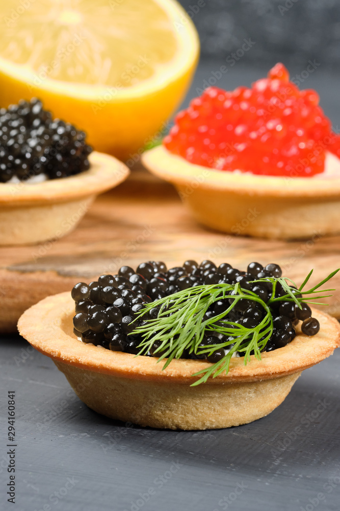 tartlets with red and black caviar on a wooden board with lemon and herbs on a gray background. Close-up tartlets with red and black caviar on a wooden board with lemon and herbs on a gray background. Close-up
