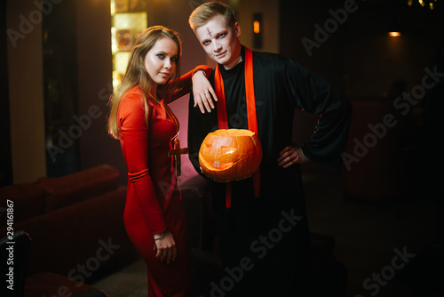 Young man in a Halloween priest costume holds a carved pumpkin in his hand. A girl in a red dress looks at the camera with a guy