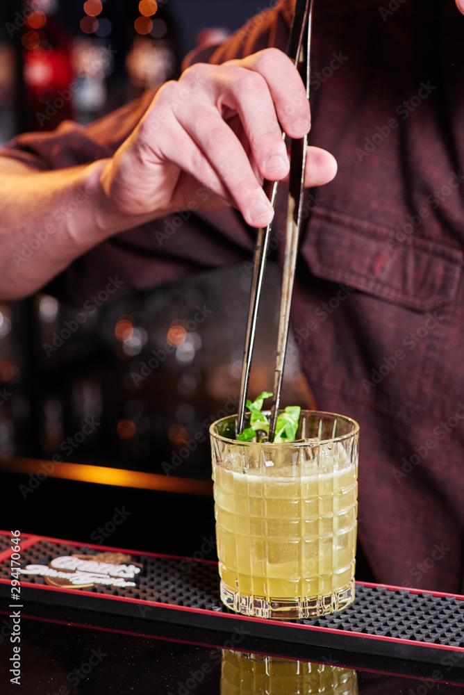 Bartender does beautiful yellow cocktail with ice, decorated pak choi  cabbage in faceted glass behind the bar. Blurred background with alcohol.  Close-up. Space. Series of photos Stock Photo | Adobe Stock