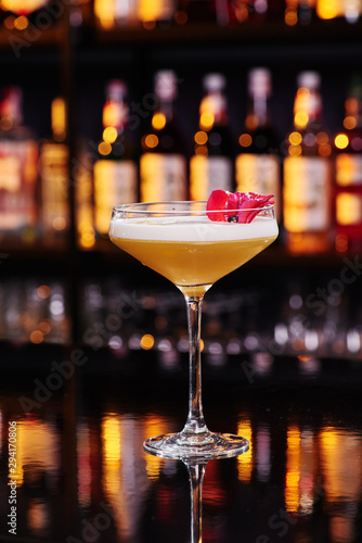 Elegant female yellow cocktail with white foam, decorated with pink flower petal on dark table against the background of blurred bar with expensive alcohol. Close-up. Space for inscriptions