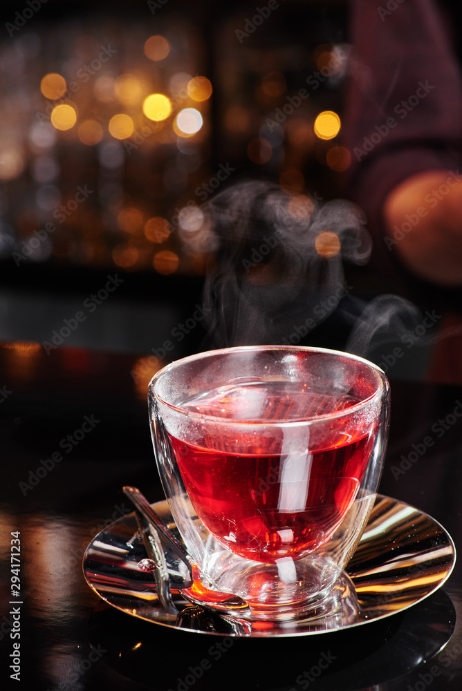 Fototapeta premium Beautiful transparent cup with red fruit tea on a shiny dark table on a blurred bar background. Close-up. Space