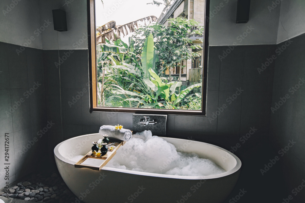 Bath with foam is filling with water, beauty products on bathtub. - obrazy, fototapety, plakaty 