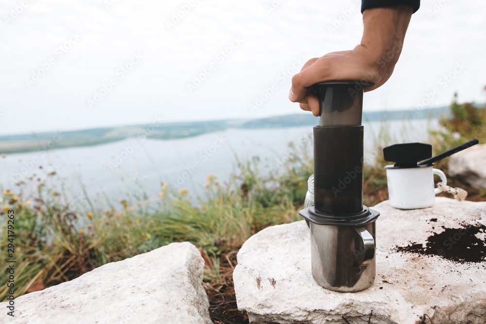 Obraz premium Traveler pressing aeropress on metal mug on cliff at lake, brewing alternative coffee at camping. Making hot drink at picnic outdoors. Trekking and hiking in mountains