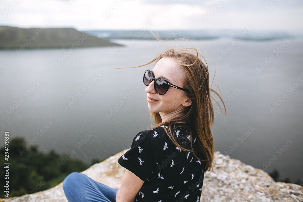 Happy girl in sunglasses sitting on top of rock mountain with beautiful view on river. Young tourist woman with windy hair relaxing on cliff. Summer vacation. Copy space Happy girl in sunglasses sitting on top of rock mountain with beautiful view on river. Young tourist woman with windy hair relaxing on cliff. Summer vacation. Copy space
