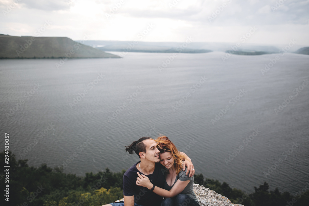 Happy hipster couple gently embracing on top of rock mountain with beautiful view on river. Tourist couple hugging on windy cliff, tranquil moment. Travel and wanderlust. Copy space Happy hipster couple gently embracing on top of rock mountain with beautiful view on river. Tourist couple hugging on windy cliff, tranquil moment. Travel and wanderlust. Copy space