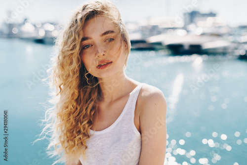 Wonderful young girl with blond curly hair in a white top on the background of the sea smiles cute and looks away