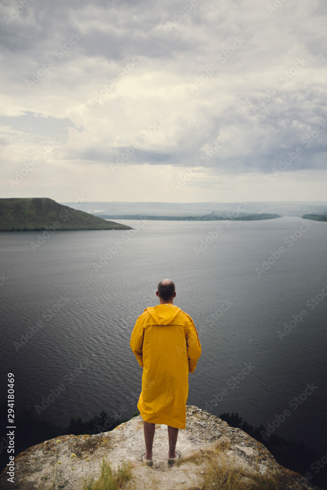 Traveler in yellow raincoat standing on cliff and looking at lake in rainy windy day. Wanderlust and travel concept. Hipster man hiking in Norway on foggy day. Atmospheric moment