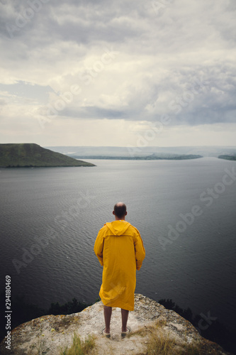 Traveler in yellow raincoat standing on cliff and looking at lake in rainy windy day. Wanderlust and travel concept. Hipster man hiking in Norway on foggy day. Atmospheric moment