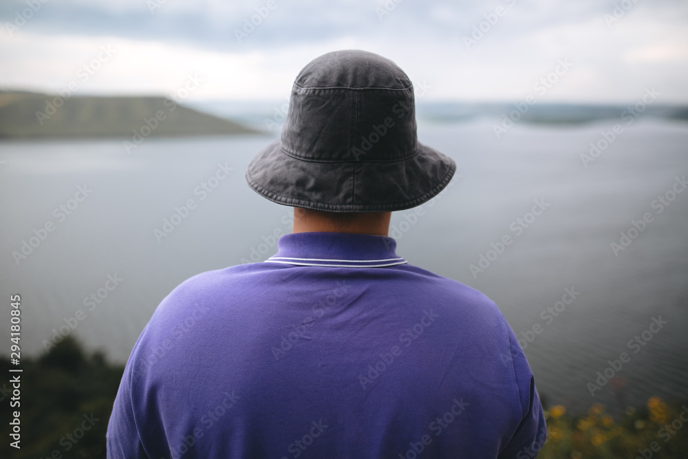 Fototapeta premium Hipster traveler in purple shirt and bucket hat standing on top of rock mountain with amazing view on river. Young camper guy exploring and traveling, back view. Copy space