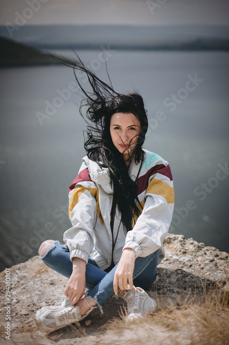 Stylish hipster girl with windy hair sitting on top of rock mountain with beautiful view on river. Young tourist woman relaxing on cliff. Atmospheric moment. Copy space