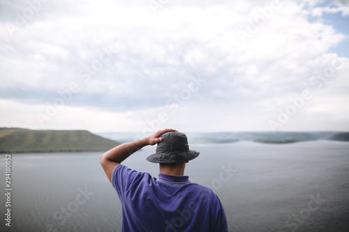 Hipster traveler in purple shirt  and bucket hat standing on top of rock mountain with amazing view on river. Young camper guy exploring and traveling, back view. Copy space