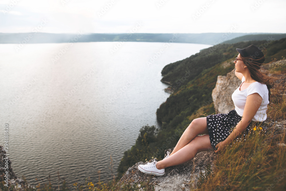 Traveler hipster girl in hat and windy hair sitting on top of rock mountain with wildflowers, enjoying beautiful sunset on river. Calm tranquil moment. Copy space