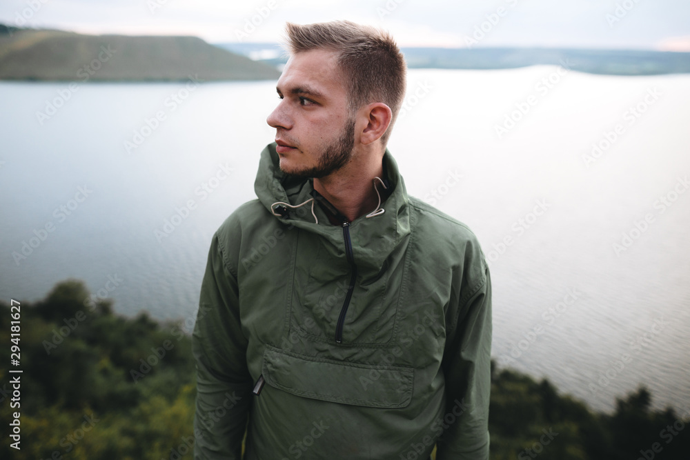 Hipster traveler standing on top of rock mountain and enjoying amazing view on river. Stylish guy in green windbreaker exploring and traveling. Atmospheric tranquil moment. Copy space Hipster traveler standing on top of rock mountain and enjoying amazing view on river. Stylish guy in green windbreaker exploring and traveling. Atmospheric tranquil moment. Copy space