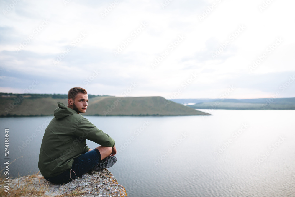 Hipster traveler sitting on top of rock mountain and enjoying amazing view on river. Stylish guy exploring and traveling. Atmospheric tranquil moment. Copy space Hipster traveler sitting on top of rock mountain and enjoying amazing view on river. Stylish guy exploring and traveling. Atmospheric tranquil moment. Copy space