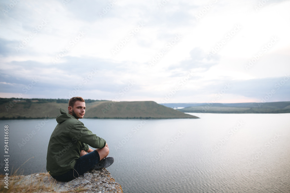 Hipster traveler sitting on top of rock mountain and enjoying amazing view on river. Stylish guy exploring and traveling. Atmospheric tranquil moment. Copy space