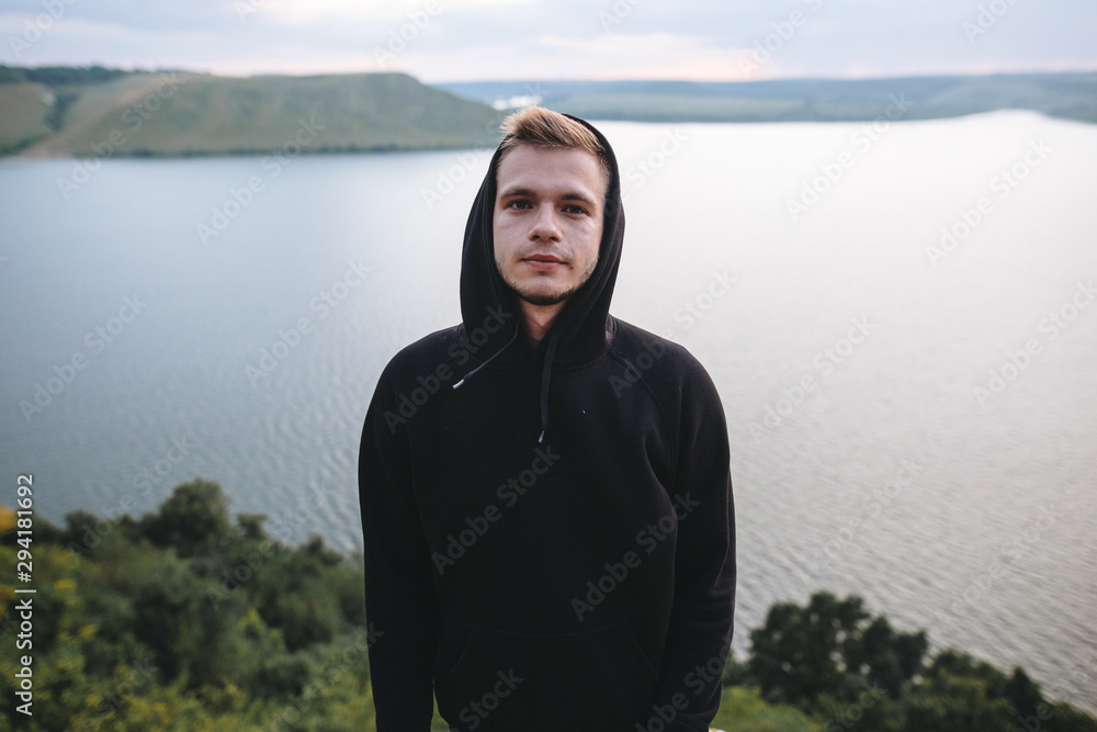 Fototapeta premium Portrait of hipster traveler in windbreaker on top of rock mountain with amazing view on river. Stylish guy exploring and traveling. Atmospheric moment. Copy space