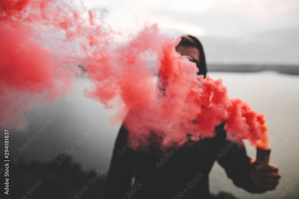 Red smoke bomb. Blurred image of ultras hooligan holding  smoke bomb in hand, standing on top of rock mountain with amazing view on river. Atmospheric moment. Copy space - obrazy, fototapety, plakaty 