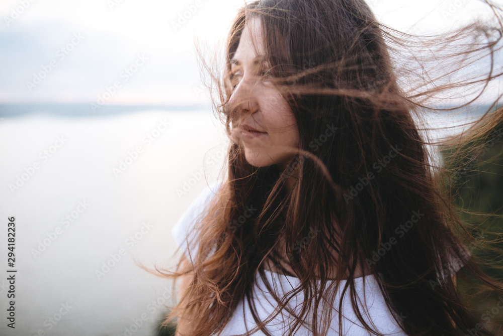 Portrait of hipster girl with windy hair standing on top of rock mountain with beautiful sunset view on river. Atmospheric tranquil and calm moment. Copy space.