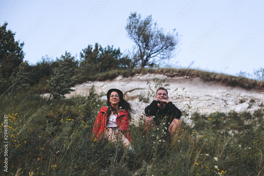 Hipster couple sitting on top of rock mountain in grass and enjoying amazing view on river in sunset. Stylish travelers exploring and traveling. Atmospheric tranquil moment. Copy space