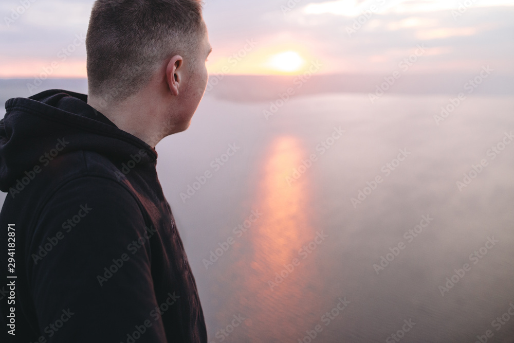 Hipster traveler enjoying amazing sunset view on river while standing on top of rock mountain. Brutal guy in windbreaker exploring and traveling. Atmospheric moment. Copy space