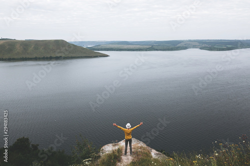 Hipster teenager in yellow pullover standing on top of rock mountain and raising hands up. Young stylish guy exploring and traveling. Atmospheric moment. Copy space