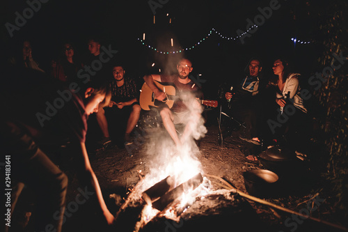 Fotografia Hipster man playing on acoustic guitar and singing song with friends travelers at big bonfire at night camp in the forest