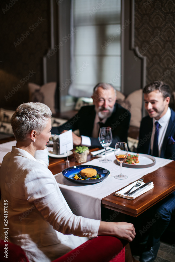Group of generous friends enjoy dinner in beautiful restaurant, close up of blonde woman sitting with back to camera at table with delicious meal, right side shot Group of generous friends enjoy dinner in beautiful restaurant, close up of blonde woman sitting with back to camera at table with delicious meal, right side shot